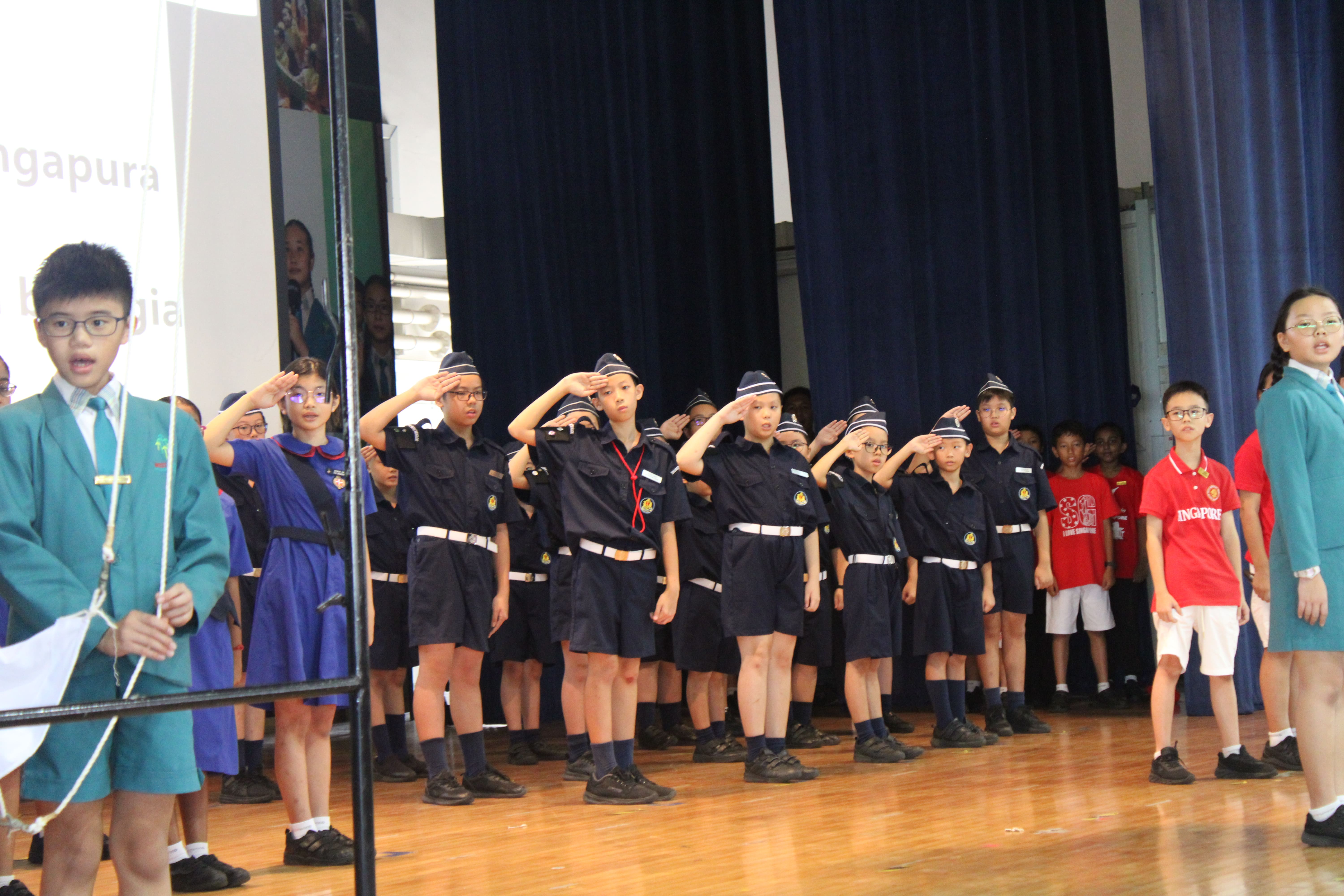 Children in dark navy uniforms and caps salute on a wooden stage during a school ceremony.